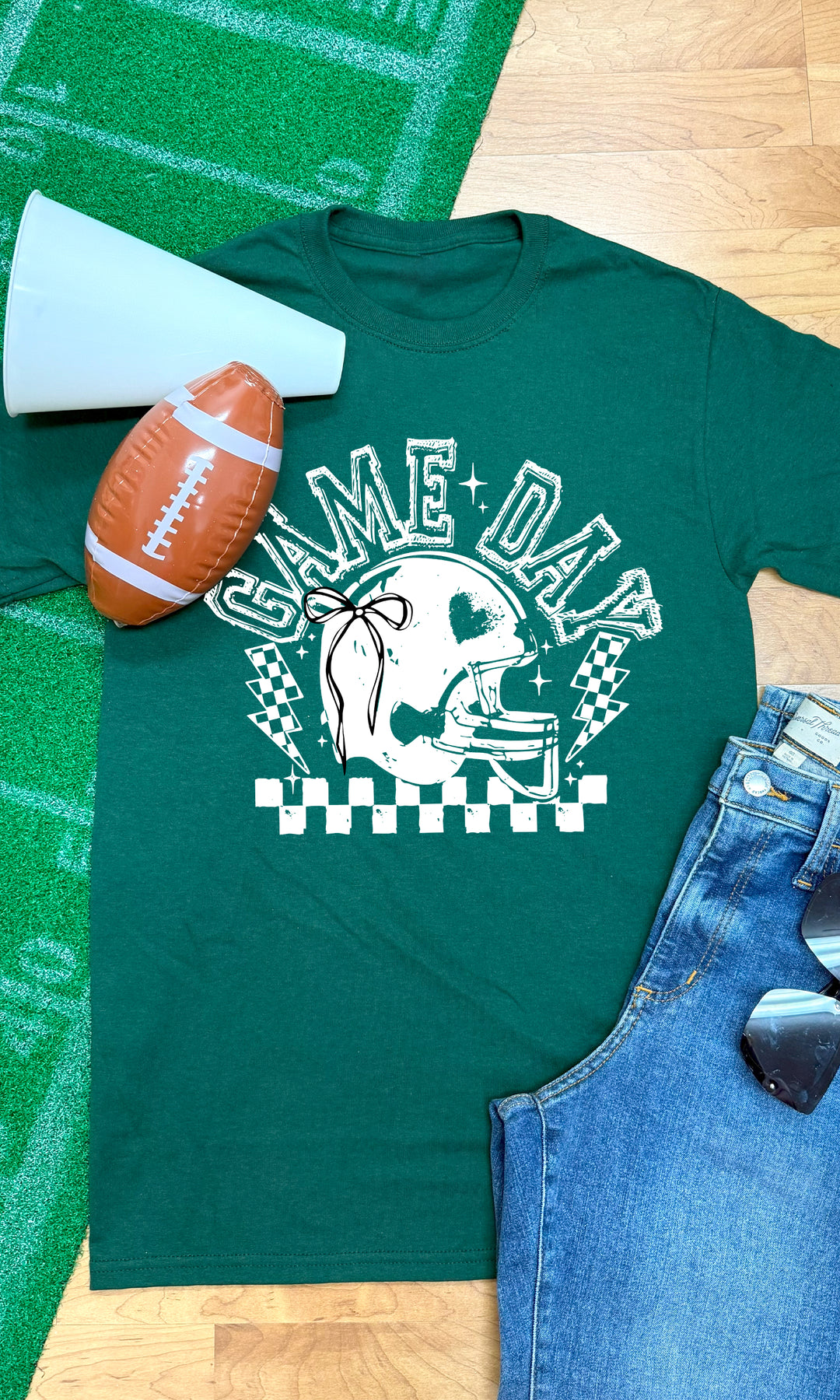 Green Game Day Football Bow Graphic T-Shirt displayed with football, megaphone, and denim shorts for a perfect game day look.