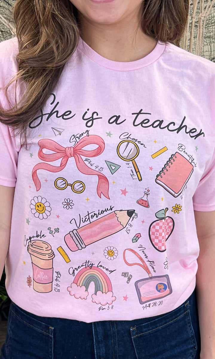 Bright pink “She is a Teacher Graphic Shirt” featuring fun school-themed illustrations, perfect for educators.